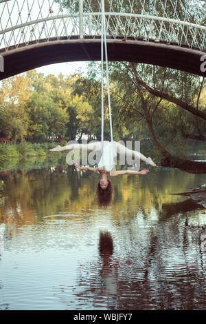 A young acrobat does twine on the canvases upside down. She hangs over the river. Stock Photo