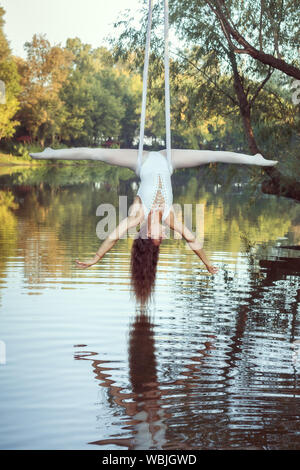 Young girl acrobat hangs on canvases upside down over the river and does the twine. Stock Photo