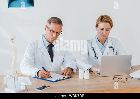 doctor writing diagnosis near coworker using laptop in hospital Stock Photo