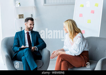handsome recruiter holding clipboard and looking at blonde employee sitting in armchair Stock Photo