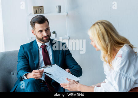 selective focus of handsome recruiter holding clipboard and pen while sitting in armchair near blonde employee Stock Photo