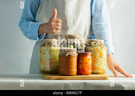 Fermented or canned vegetables, different in jars stand on the salt against the background of a woman in an apron. Processing the autumn harvest. Natu Stock Photo