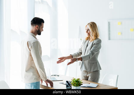 happy blonde recruiter gesturing in office near handsome bearded man Stock Photo
