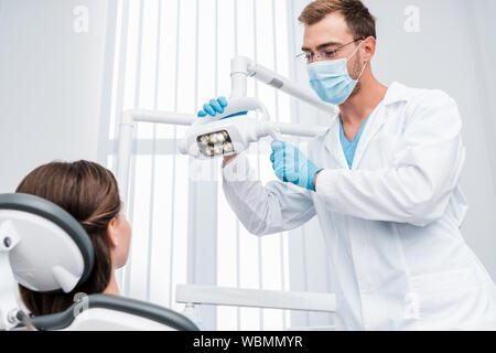 selective focus of dentist in medical mask and blue latex gloves touching medical lamp near patient Stock Photo