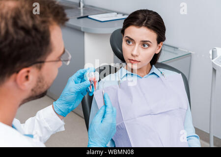 selective focus of young woman looking at dentist in latex gloves holding tooth model and dental instrument Stock Photo