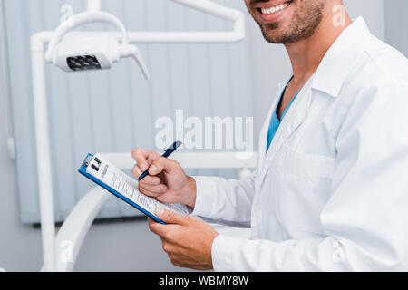 cropped view of bearded man in white coat holding pen and clipboard in dental clinic Stock Photo