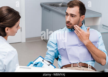 selective focus of upset man gesturing and looking at attractive dentist in latex gloves holding pen and clipboard Stock Photo