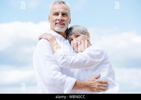 happy senior couple in white shirts embracing under blue sky Stock Photo