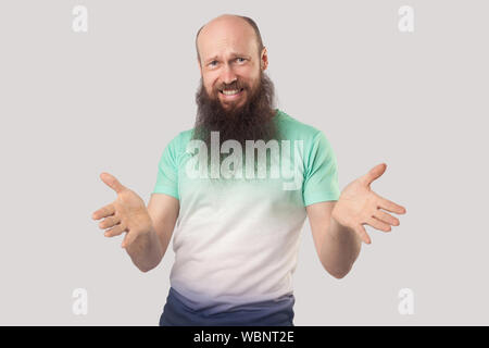 What do you need? Portrait of angry middle aged bald man with long beard in light green t-shirt standing, looking at camera with raised arms and askin Stock Photo
