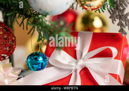 Close up a red gift box tied with a white ribbon placed under the pine tree. A blue small bell and snowflke hanging on the branch with blurred backgro Stock Photo
