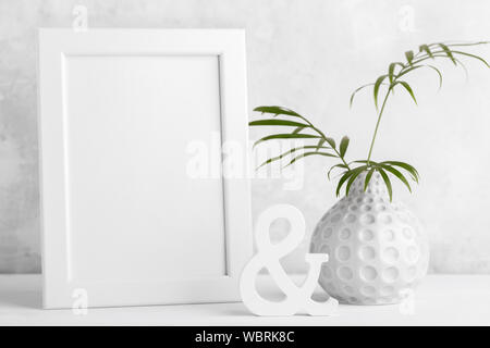 Small modern vase with branch of palm leaves and empty frame mock up on the shelf, grey stone wall room minimalistic interior design Stock Photo