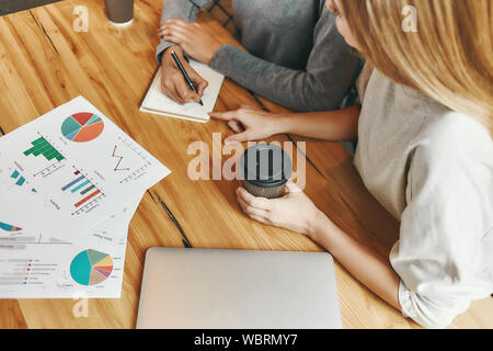 Cropped shot of two female colleagues. They are sharing new ideas with each other and making notes as they sit in a cafeteria drinking coffee. Charts, diagrams and laptop on the table Stock Photo