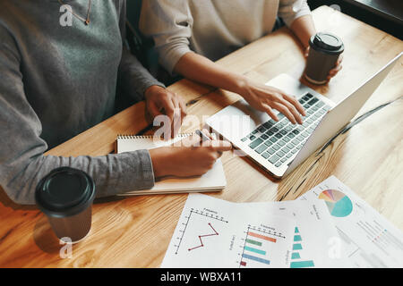 Cropped shot of two female colleagues. They are typing on a laptop and making notes as they sit in a cafeteria enjoying a cup of coffee. Charts and diagrams on the table. Horizontal shot Stock Photo
