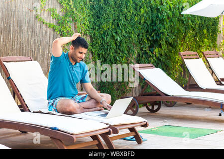Portrait of clever handsome bearded young adult freelancer man in blue t-shirt and shorts sitting on cozy deck chair with laptop on poolside, working Stock Photo
