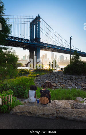 New York City young people, rear view of a young couple sitting in John Street Park Brooklyn watching the sun setting behind the Manhattan Bridge, NYC Stock Photo
