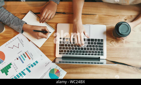 Top view of female hands of two colleagues. They are typing on a laptop and making notes as they sit in a cafeteria enjoying a cup of coffee. Charts and diagrams on the table. Horizontal shot Stock Photo