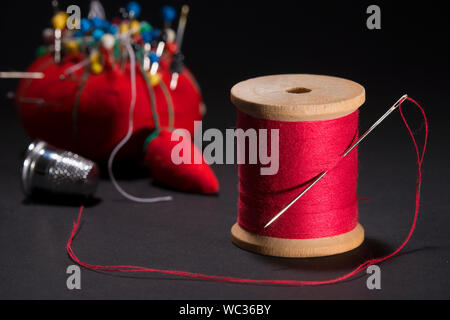 Close up of sewing tools.  Wooden spool of red thread with threaded needle inserted between threads.  Pin cushion and thimble in out of focus backgrou Stock Photo