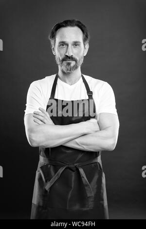 Confident in his culinary craft. Cook with beard and mustache wearing apron red background. Man mature cook posing cooking apron. Chief cook and professional culinary. Cook food at home. Fine recipe. Stock Photo