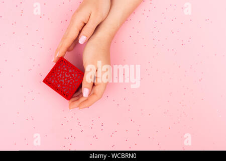The girl takes a red patterned box with a ring with two hands. Top view on a pink background with place for text. Glitter in the background. Stock Photo