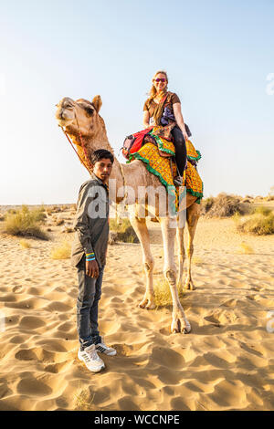 A woman smiling atop a camel she is riding with her camel driver / guide standing below, Thar desert, Rajasthan, India. Stock Photo