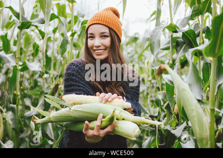 Beautiful carefree long hair asian girl in the yellow hat and knitted sweater holding cobs of corn in the autumn field. Sensitivity to nature concept