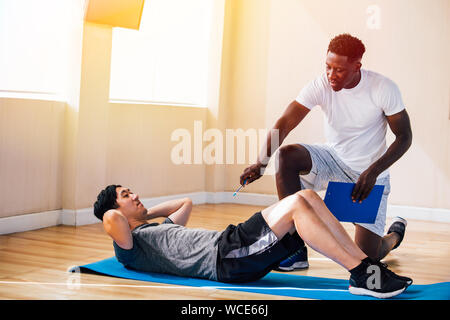 Side view of Asian man lying on floor doing abdominal exercises while African American trainer kneeling besides and teaching in fitness club Stock Photo