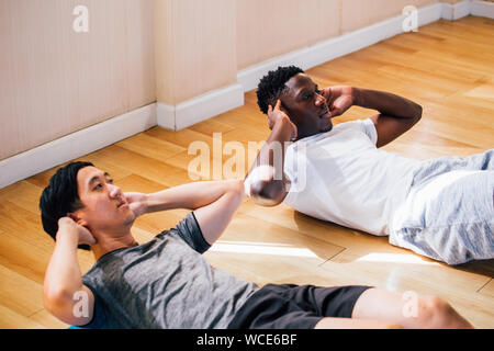 From above multiethnic athletic men lying on floor holding hands behind head doing and abdominal exercises in fitness club on daytime Stock Photo