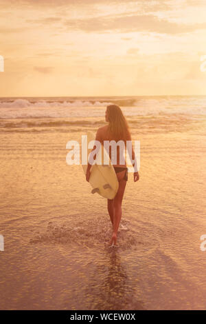 A young woman in bikini stands with her back to the camera and holds a surfboard by the ocean. Warm tinting. Surf concept. Full-length photo Stock Photo