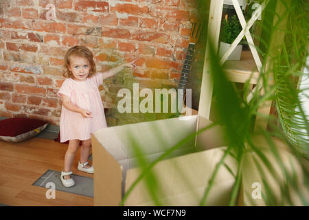 New life. Cute little girl moved to a new house or apartment. Look happy and confident. Moving, relations, lifestyle concept. Discovering her new kids room, pointing on the brick wall, smiling. Stock Photo