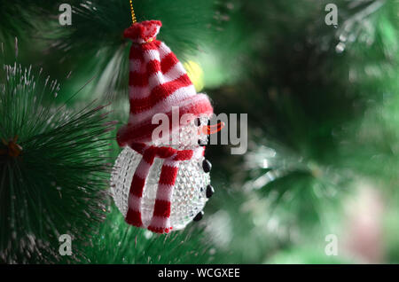 Toy snowman in a beautiful knitted red hat and scarf hanging on a green Christmas tree Stock Photo
