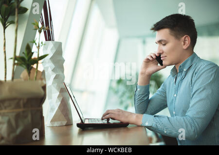 young handsome businessman has conversation on his smartphone and works in his laptop while waiting for lunch in cafe near office. Stock Photo