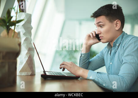 young handsome surprised businessman has conversation on his smartphone and works in his laptop while waiting for lunch in cafe near office. Stock Photo