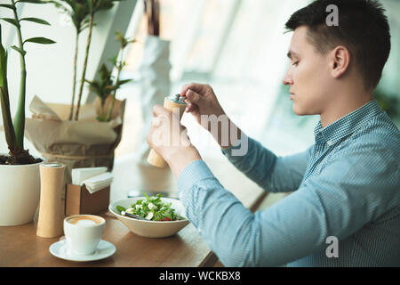 young handsome man salting salad preparing to eat his lunch and drinks coffee during break at cafe near office. Stock Photo
