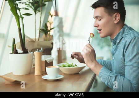 young handsome man eats salad and drinks coffee for lunch during break at cafe near office looks hungry. Stock Photo