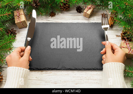 Perspective view girl holds fork and knife in hand and is ready to eat. Empty black slate rectangular plate on wooden christmas background. holiday di Stock Photo