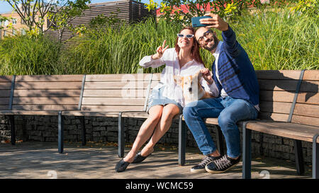 The happy couple are sitting on the bench in the park with little dog and do selfie portrait. Cute corgi Pembroke puppy on its owners hands. Concepts