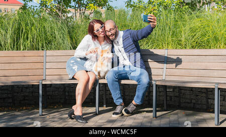 The happy couple are sitting on the bench in the park with little dog and do selfie portrait. Cute corgi Pembroke puppy on its owners hands. Concepts Stock Photo