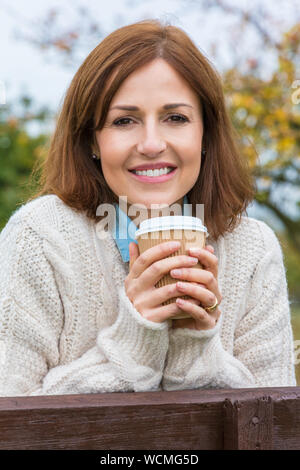 Attractive happy thoughtful middle aged woman leaning resting on fence in the countryside smiling with a cup of takeout tea or coffee Stock Photo