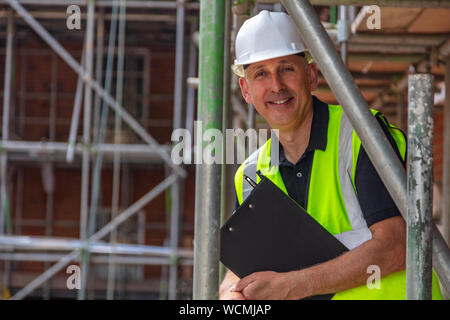 Male builder foreman, construction worker, surveyor or site manager holding a clipboard, wearing a white hard hat and hi vis vest and smiling Stock Photo