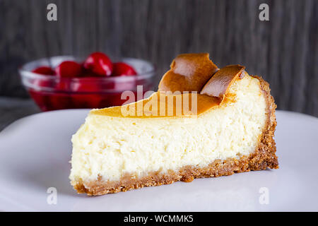 A slice of New York style cheesecake with a bowl of cherry sauce blurred in the background.  White plate.  Neutral background. Stock Photo