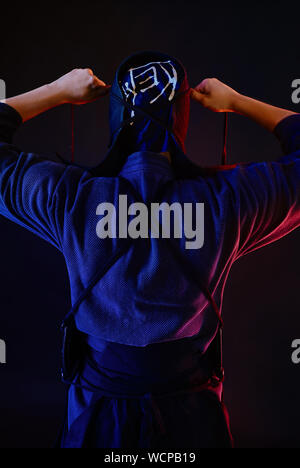 Close up shot, Kendo fighter wearing in an armor and traditional kimono is tying the lacing on his helmet standing back against a black background. Stock Photo