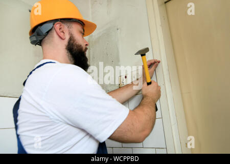 Construction worker and handyman working on renovation work. Builder with yellow hammer strikes and nails a nail into wooden wall of kitchen door. Stock Photo