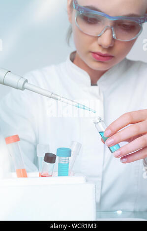 Young female scientist loads liquid sample into disposable plastic vial, scientific background with copy-space, shallow DOF, focus on the vial and pip Stock Photo