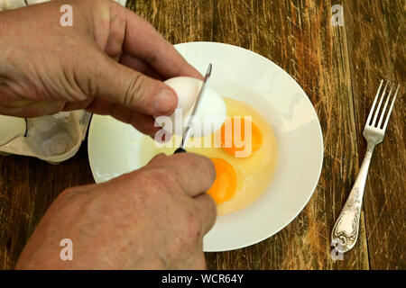 Homemade kitchen. Cooks hands break white chicken eggs with a table knife in a white plate. Close-up. Stock Photo