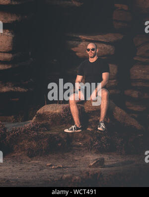 Young confident muscular male sat looking at the camera in front of a rocky structure Stock Photo