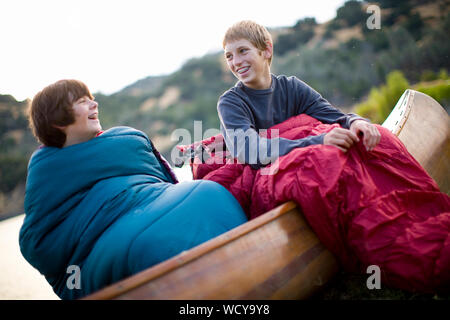Two smiling boys sitting in a wooden canoe inside sleeping bags. Stock Photo