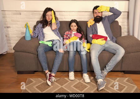 Tired parents and kid. Cleaning all day exhausting occupation. Exhausting cleaning day. Family mom dad and daughter with cleaning supplies sit on couch. Family care about cleanliness. Finish cleaning. Stock Photo