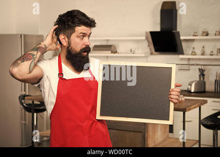 What to cook. urprise. This is what you need. Hipster in kitchen. Restaurant or cafe menu. Advertisement. Bearded man in red apron. Mature male. Bearded man cook. Man chef with board, copy space. Stock Photo