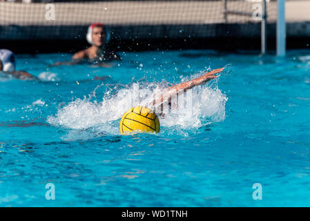 Floating water polo ball and emerging arm from submerged player while goalie looks on during competition match. Stock Photo