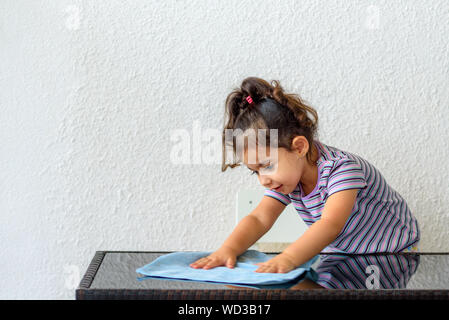 Cute brunette child helping her mom washing modern design garden furniture, glass table on home terrace. Little girl make cleaning with water and blue towel. Stock Photo